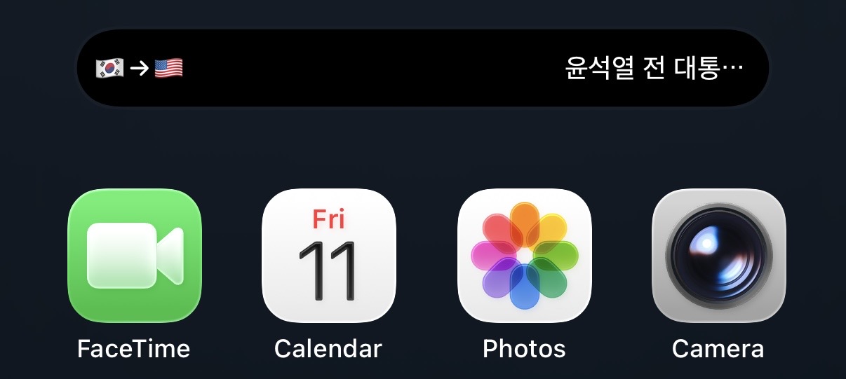 The compact Dynamic Island on the home screen above the FaceTime, Calendar, Photos, and Camera icons: a Korean flag, an arrow, and a US flag at the left, and the start of the Korean line being read at the right
